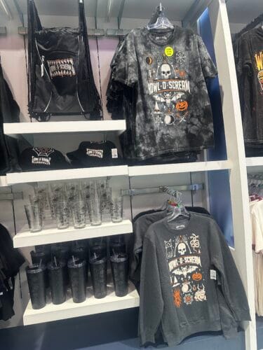 Gray Howl-O-Scream shirt and sweatshirt with Halloween symbols beside bags, caps, clear glasses, and black tumblers.
