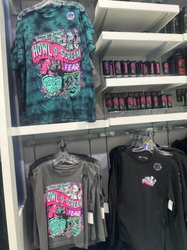 Teal tie-dye and gray From the Deep graphic T-shirts beside black Howl-O-Scream apparel and souvenir glasses.